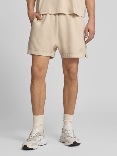Mężczyzna nosi Regular Fit Sweatshorts mit Modal und Baumwoll-Anteil od adidas Sportswear, efekt: jednolity kolor, detal z logo, elastyczny ściągacz z tunelem, wpuszczane kieszenie, mieszanka materiałów z domieszką modalu i bawełny, jasny beż, tekstura gładka, Ganzkörperansicht z przodu.