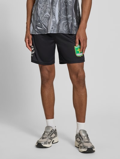 Männermodel trägt Relaxed Fit Shorts mit Logo-Print von COLUCCI, schwarzer Stoff, elastischer Bund, Motiv-Stitching, Innenbeinlänge 18 cm, Ganzkörperansicht von vorne.
