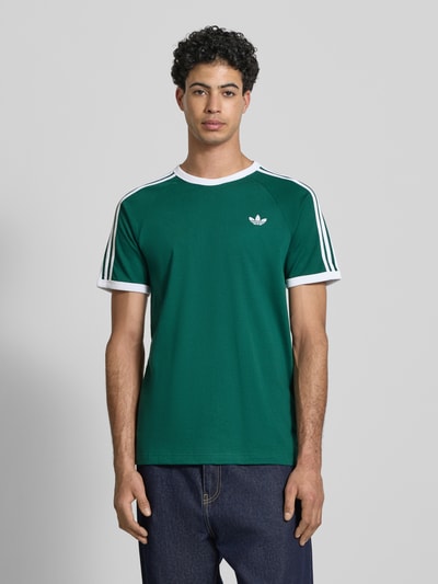 Męski model nosi T-Shirt mit Label-Stitching od adidas Originals, bawełniany materiał, krój regular fit, ściągacze, kolor zielony, widok z przodu na całe ciało.