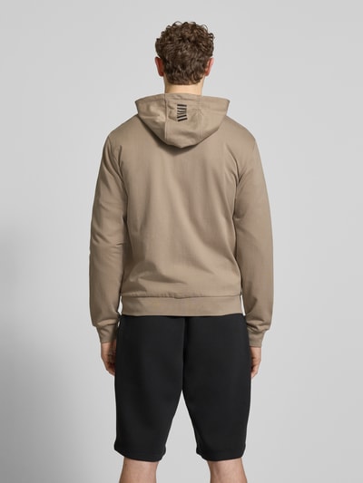 Męski model nosi Slim Fit Sweatjacke mit Kapuze od EA7 Emporio Armani, beżowy bawełniany materiał, logo z tyłu, krój slim fit, bawełniany miks, kaptur ze ściągaczem, zamek dwukierunkowy, kieszenie boczne, elastyczne wykończenia, długość tyłu w rozmiarze M: 69 cm, długość rękawa w rozmiarze M: 69 cm, pełny widok sylwetki z tyłu.