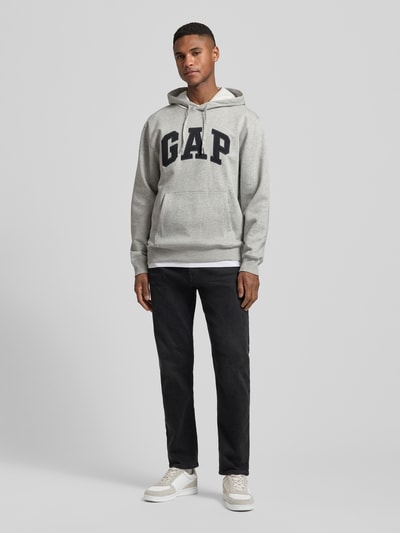 Männermodel trägt Hoodie mit Logo und Känguru-Tasche von GAP in Grau, Baumwoll-Mix, Regular Fit, schwarze Hose, Ganzkörperansicht von vorne.