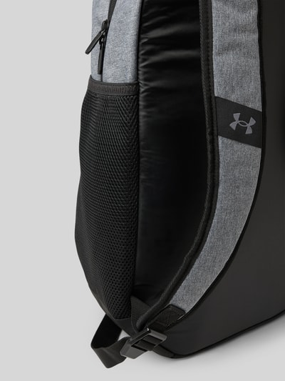 Produktfoto, Rugzakfunctie met draagband en verstelbare schouderband van Under Armour, hoofvak met ritssluiting, labelprint, zijvak met ritssluiting, grijs en zwart, breedte 31 cm, lengte 48 cm, diepte 18 cm, bandlengte 53 cm.