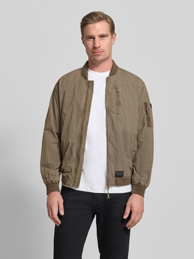 Männermodel trägt khakifarbenen Blouson mit Label-Patches Modell 'IBE' von khujo mit Reißverschluss, Brusttasche, Reißverschlusstasche am Ärmel, Seitliche Eingrifftaschen, Stehkragen und Gerader-Schnitt, Ganzkörperansicht von vorne.