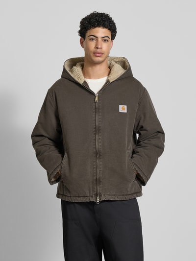 Männermodel trägt Jacke mit Zweiwege-Reißverschluss Modell 'MITCH' von Carhartt aus brauner Baumwolle mit Kapuze und Label-Detail, Ganzkörperansicht von vorne.