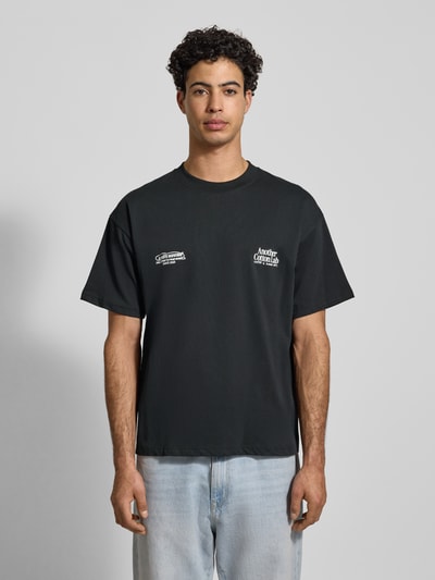 Männermodel trägt T-Shirt mit Label-Print von ANOTHER COTTON LAB aus reiner Baumwolle, lockerer Schnitt, gerippter Rundhalsausschnitt, Ganzkörperansicht von vorne.