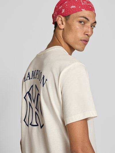 Männermodel trägt Relaxed Fit T-Shirt aus reiner Baumwolle von Champion in Creme mit blauem Statement-Print, Rückenansicht, Nahaufnahme Oberkörper. Herren T-Shirt, Label-Stitching erkennbar, Rücklänge bei Größe M 70 cm.