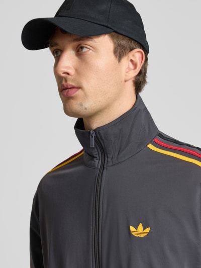 Männermodel trägt Regular Fit Trainingsjacke mit mehrfarbigen Kontraststreifen Modell 'FIREBIRD TT' von adidas Originals, Logo-Stitching, unifarbene Optik, Stehkragen, langarm, Nahaufnahme Oberkörper.