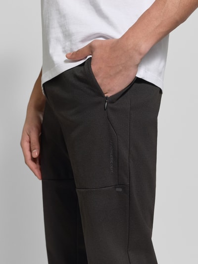 Mężczyzna nosi spodnie dresowe z elastycznej mieszanki materiałów z domieszką bawełny model 'GORDON' od Jack & Jones, krój slim fit, elastyczny pas z tunelem, elastyczne zakończenia nogawek, boczne kieszenie zapinane na zamek błyskawiczny, długość wewnętrzna nogawki w rozmiarze m: 73 cm, ciemny kolor, prosty materiał, gładka tekstura, zbliżenie na górną część nogawki