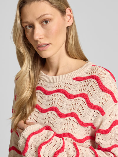 Frauenmodel trägt Regular Fit Pullover in Strick-Optik Modell 'Wave' von Vila aus reiner Baumwolle mit Lochmuster, Rundhalsausschnitt, beige mit roten Wellenlinien, Nahaufnahme Oberkörper