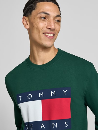 Männermodel trägt Regular Fit Sweatshirt aus reiner Baumwolle von Tommy Jeans in Dunkelgrün mit großem Logo-Print vorne, Nahaufnahme Oberkörper. Herren Sweatshirt von Tommy Jeans.