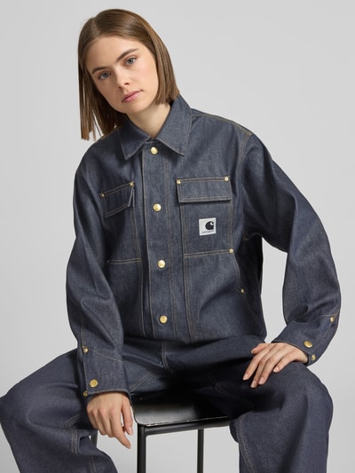 Frauenmodel trägt Regular Fit Jeansjacke mit Logo-Patch Modell 'Arctic' von Carhartt Work In Progress aus reiner Baumwolle in Dunkelblau, goldene Knöpfe, Umlegekragen, Brust- und Eingrifftaschen, Nahaufnahme Oberkörper.