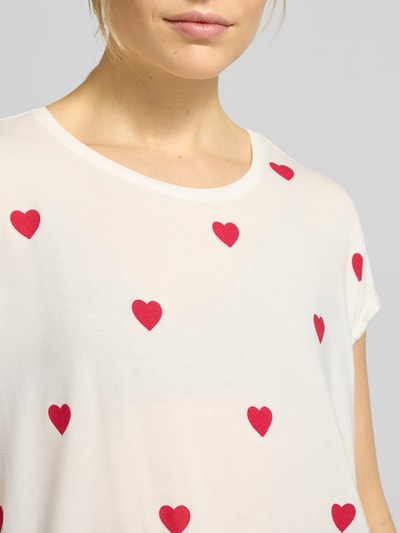 Frauenmodel trägt Relaxed Fit T-Shirt aus Lyocell-Mix Modell 'AVA' von Vero Moda mit rotem Allover-Herzprint auf weißem Grund, Nahaufnahme Oberkörper.
