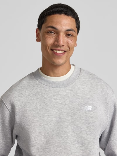 Männermodel trägt Sweatshirt mit Rundhalsausschnitt und Label-Stitching von New Balance aus grauem Baumwoll-Mix, Nahaufnahme Oberkörper. Relaxed-Fit, Rückenlänge bei Größe M 67 cm, Ärmellänge 63 cm.