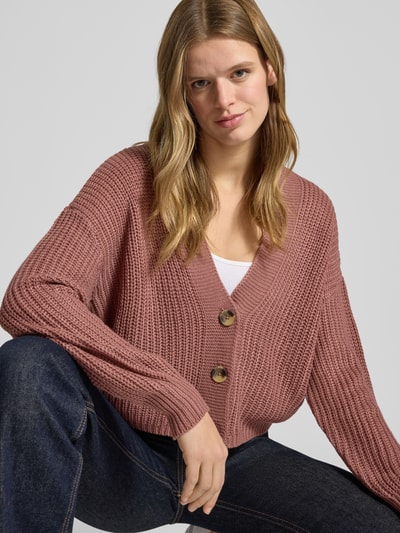 Frauenmodel trägt Regular Fit Strickjacke mit einreihiger Knopfleiste Modell 'CAROL NICE' von Only, rosafarbene Strickoptik, tiefer V-Ausschnitt, langer Ärmel, Cropped-Länge, Nahaufnahme Oberkörper.