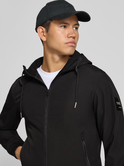 Männermodel trägt Regular Fit Jacke mit Logo-Detail von JACK & JONES in Schwarz mit Kapuze und Reißverschlusstaschen, Nahaufnahme Oberkörper. Herren Jacke, Softshell, elastische Abschlüsse, Rückenlänge 66 cm, Schulterbreite 46 cm, Brustweite 53 cm, Ärmellänge 67 cm.