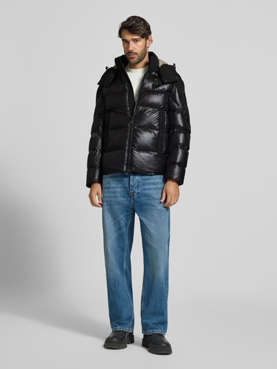 Męski model nosi czarną, błyszczącą Steppjacke mit Zweiwege-Reißverschluss Modell 'KIERNAN' von Blauer USA, krój regular fit, z kapturem, label-detail, szerokość w rozmiarze M: 59 cm, długość tyłu: 67 cm, szerokość ramion: 49 cm, długość rękawa: 65 cm; cała sylwetka z przodu.
