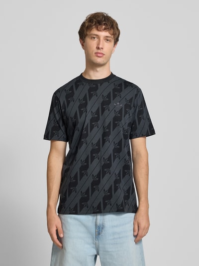 Männermodel trägt T-Shirt aus reiner Baumwolle Modell 'MONOGRAMM AOP' von adidas Originals in Schwarz mit Muster, Ganzkörperansicht von vorne.