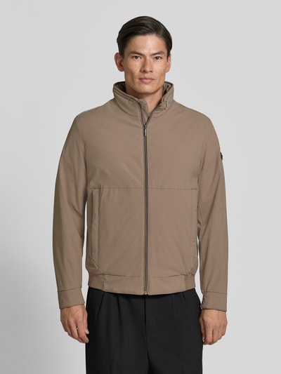 Männermodel trägt Regular Fit Jacke mit stójka von bugatti w beżowym kolorze, wpuszczane kieszenie, zamek błyskawiczny, elastyczny pas, długość tyłu w rozmiarze 50: 67 cm, szerokość ramion w rozmiarze 50: 45 cm, szerokość klatki piersiowej w rozmiarze 50: 58 cm, długość rękawów w rozmiarze 50: 67 cm, całkowity widok z przodu