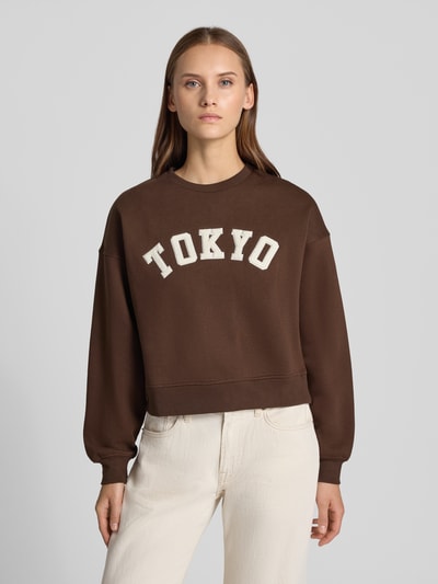 Vrouwenmodel draagt Relaxed Fit Sweatshirt aus Baumwoll-Mix Model 'Tatum' van JJXX in donkerbruin met statementpatch, katoenmix met ribboordjes, dames sweatshirt, met lange mouwen, ronde hals, schouderpartijen, effen design, vooraanzicht bovenlichaam.