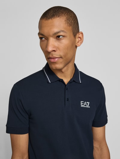 Męski model nosi Regular Fit Poloshirt mit Logo-Print von EA7 Emporio Armani, granatowa bawełna, kołnierzyk polo, krótki rękaw, elastyczne mankiety, zbliżenie na górną część ciała.