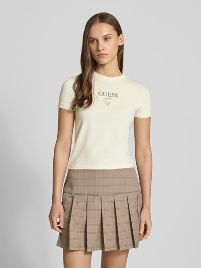Frauenmodel trägt T-Shirt mit Logo und Rundhalsausschnitt von Guess Jeans in Creme, Slim Fit, elastischer Baumwollmix, Logo-Print, Ganzkörperansicht von vorne.