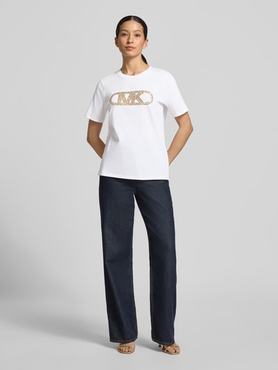 Frauenmodel trägt T-Shirt mit Logo und Rundhalsausschnitt von MICHAEL Michael Kors, Reine Baumwolle, Regular-Fit, weiß mit goldenem Logo, Rückenlänge bei Größe S: 62 cm, Ganzkörperansicht von vorne.