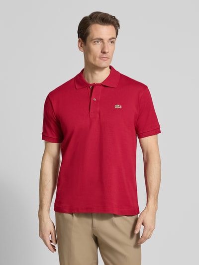 Męski model nosi Regular Fit Poloshirt aus reiner Baumwolle von Lacoste, jednolity kolor, krój regular fit, wyhaftowane logo, kołnierzyk polo, krótki rękaw, prążkowane wykończenie rękawów, rozcięcia po bokach, pika, bawełna, całkowity widok z przodu.
