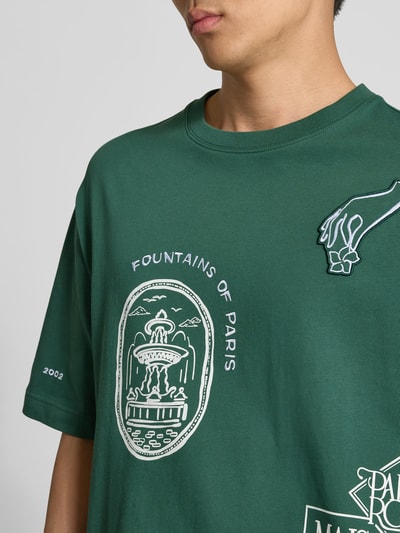 Männermodel trägt Oversized T-Shirt aus Bio-Baumwolle von MAISON KITSUNÉ in Dunkelgrün mit weißem Label-Print, Kurzärmelig, Nahaufnahme Oberkörper.