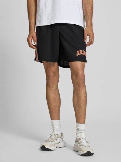 Männermodel trägt Regular Fit Shorts mit Logo-Print von KARL KANI, Schwarz mit Strukturmuster, elastischer Bund, Nahaufnahme Unterkörper, Herren Shorts, Bundweite Größe M: 37 cm, Innenbeinlänge: 19 cm, Vorderansicht.