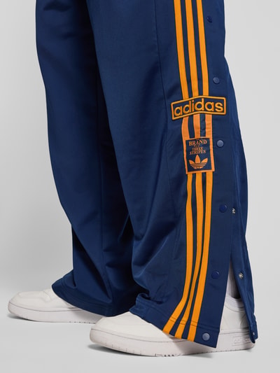 Männermodel trägt Sweatpants mit elastischem Bund von adidas Originals in Dunkelblau mit orangenen Streifen, Label-Stitching, Eingrifftaschen, Lockerer Schnitt, Innenbeinlänge bei Größe M 75 cm, Nahaufnahme Beinansicht.