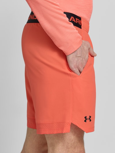 Mężczyzna nosi Straight Fit Shorts mit Logo-Print Modell 'Vanish' od Under Armour w kolorze koralowym, elastyczny pas i kieszenie, Szerokość w rozmiarze M: 38 cm, długość nogawki od wewnątrz: 15 cm, krój straight fit, widok z boku na wysokości bioder.