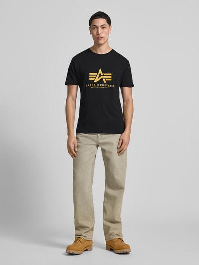 Männermodel trägt Herren T-Shirt mit Label-Detail Modell 'Basic' von Alpha Industries in Schwarz mit gelbem Print, Reine Baumwolle, Gerader-Schnitt, Rundhalsausschnitt, Rückenlänge bei Größe M: 67 cm, Ganzkörperansicht von vorne.