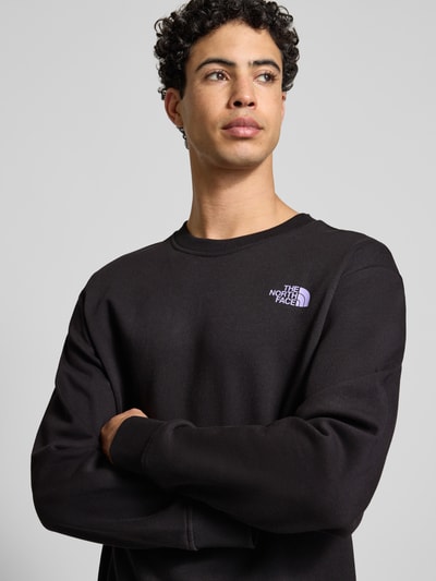 Męski model nosi Bluza z nadrukiem z logo Sweatshirt mit Label-Print od The North Face, bawełna, krój relaxed fit, prążkowane wykończenia, okrągły dekolt, czarna, półzbliżenie na górną część ciała.