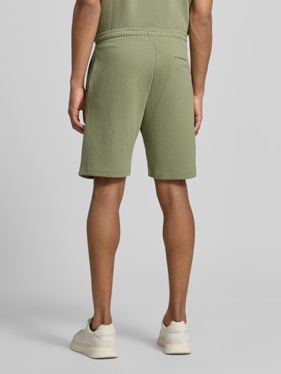 Männermodel trägt Regular Fit Shorts mit Tunnelzug von MCNEAL aus Baumwoll-Mix mit Stretch-Anteil, mit Eingrifftaschen, Gesäßtasche, Bundweite bei Größe M 43 cm, Innenbeinlänge bei Größe M 23 cm, Struktur in hellem Oliv, Ganzkörperansicht Rückansicht.
