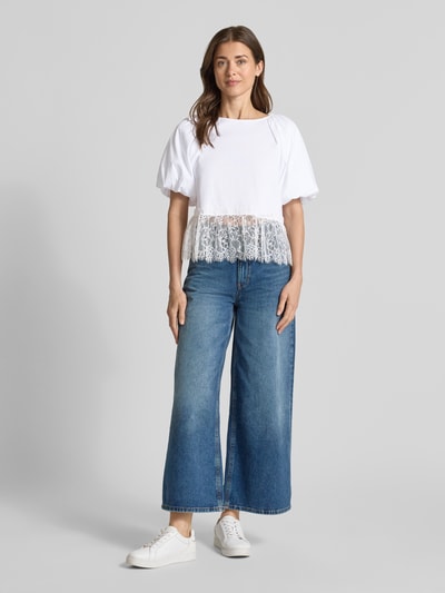 Frauenmodel trägt T-Shirt mit Spitze und Ballonärmeln von Rich & Royal aus Reine Baumwolle, Straight Fit, Rundhalsausschnitt, Rückenlänge bei Größe S: 56 cm, weiße Farbe, blaue Jeans, weiße Sneaker, Ganzkörperansicht von vorne.