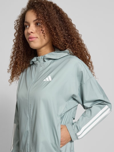 Modelka nosi Windbreaker mit Kapuze und Logo od adidas Sportswear, relaxed fit, zamek błyskawiczny, kaptur, kieszenie, miękka tekstura, logo z przodu, jasnoszary kolor, szerokość w rozmiarze S: 53 cm, długość tyłu: 58 cm, szerokość ramion: 66 cm, długość rękawa: 50 cm, zbliżenie na górną część ciała z przodu.