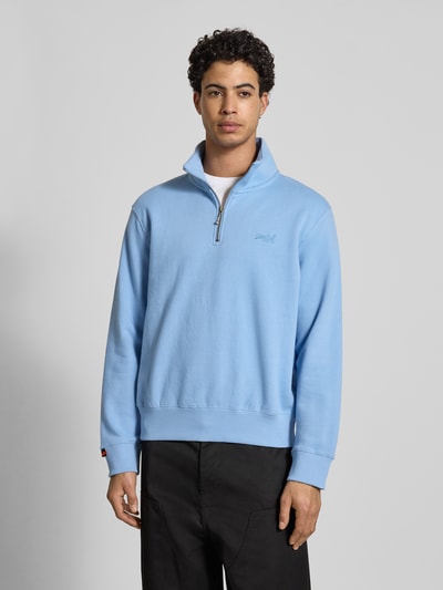 Mannelijk model draagt Regular Fit Sweatshirt met Logo-Stitching van Superdry in lichtblauw katoenmix, opstaande kraag met ritssluiting, elastische boorden, ruglengte bij maat M 65 cm, mouwlengte 63 cm, kleur effen, textuur glad, voorkant, overromp close-up.