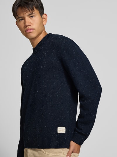 Männermodel trägt Regular Fit Strickpullover mit Rundhalsausschnitt von JACK & JONES, Strukturstrick, dunkelblau, Nahaufnahme Oberkörper. Material-Mix mit Baumwoll-Anteil, gerippte Abschlüsse, Logo-Patch sichtbar.
