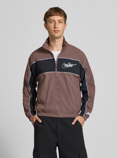 Männermodel trägt Regular-Fit Trainingsjacke mit Logo von Nike in Braun mit schwarzen und weißen Details, Reißverschluss, Stehkragen, Ganzkörperansicht von vorne. Rückenlänge 67 cm, Ärmellänge 64 cm.