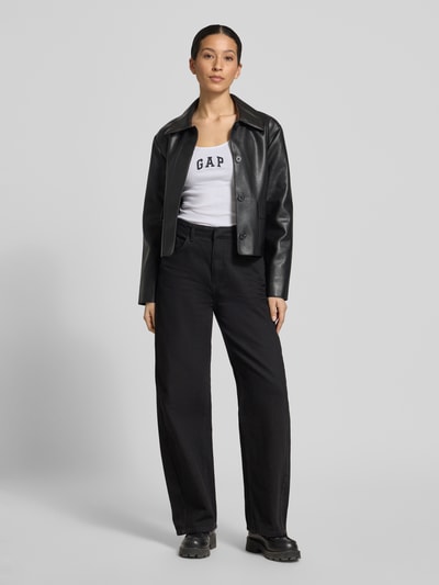 Frauenmodel trägt Slim Fit Top mit Logo-Print von GAP in Weiß, kombiniert mit schwarzer Hose und Jacke, Ganzkörperansicht von vorne. Elastischer Baumwoll-Mix, Rundhalsausschnitt, Rückenlänge 58 cm bei Größe S.