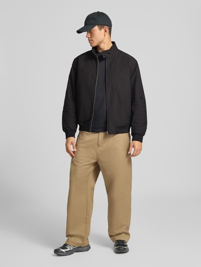 Mannelijk model draagt Regular Fit jack met logostitching Model 'HARRINGTON' van Superdry in zwarte elastische materiaalmix met opstaande kraag en ritssluiting, herenjacks, ruglengte maat M 66 cm, schouderbreedte 50 cm, borstwijdte 60 cm, mouwlengte 65 cm, kleuren zwart en beige, textuur glad, geheel lichaam van voren.