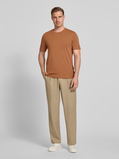Männermodel trägt T-Shirt mit Label-Detail von bugatti in Braun, gerade geschnitten, aus Baumwolle, kombiniert mit beigen Hosen, Ganzkörperansicht von vorne.