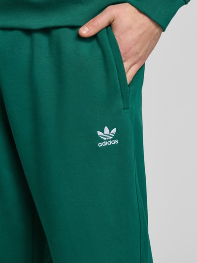 Männermodel trägt Wide Fit Sweathose mit elastischem Bund von adidas Originals in Dunkelgrün, Logo-Stitching, Nahaufnahme Hüfte.