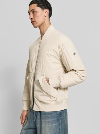 Männermodel trägt Bomberjacke mit Daunen-Feder-Füllung von Moose Knuckles in Beige mit Zweiwege-Reißverschluss, Känguru-Tasche mit Patten, lockere Passform, Kapuze mit Tunnelzug, Rückenlänge 63 cm, Schulterbreite 48 cm, Ärmellänge 65 cm, Ganzkörperansicht von vorne.