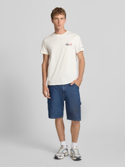 Männermodel trägt weißes Slim Fit T-Shirt aus reiner Baumwolle von Tommy Jeans, blaue Jeansshorts, Ganzkörperansicht von vorne.