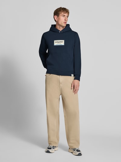 Männermodel trägt Hoodie mit Label-Print Modell 'FREDERIKSBERG' von JACK & JONES in Dunkelblau, lockerem Schnitt, Ganzkörperansicht von vorne, Baumwoll-Mix, Kängurutasche, gerippte Abschlüsse.