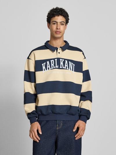Männermodel trägt Oversized Sweatshirt mit Polokragen Modell 'Heritage' von KARL KANI im Baumwoll-Mix mit Logo-Print, navy-beige, elastische Abschlüsse, Rückenlänge bei Größe M 65 cm, Oversized-Fit, Ganzkörperansicht von vorne.