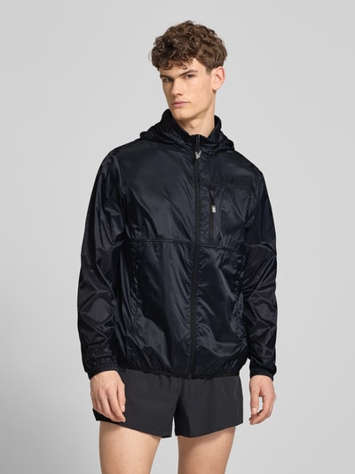 Männermodel trägt Regular Fit Jacke mit Stehkragen Modell 'Berg' von Ellesse in Schwarz, glänzende Textur, Ganzkörperansicht von vorne, Napoleontasche, Reißverschlusstaschen, elastische Abschlüsse, Regular Fit, Rückenlänge 73 cm, Schulterbreite 49 cm, Brustweite 58 cm, Ärmellänge 70 cm.