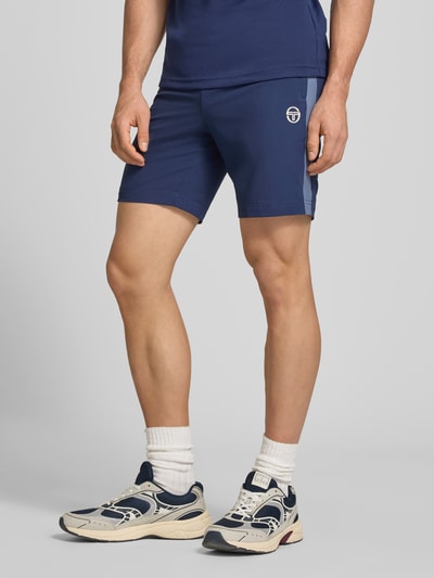 Männermodel trägt Shorts mit Logo-Print Modell 'GOCCIA' von SERGIO TACCHINI in Dunkelblau, lockerer Schnitt, Nahaufnahme Oberschenkel, Kontext: Herren Shorts, Material-Mix mit Stretch, elastischer Bund, Logo-Print, Bundweite 38 cm, Innenbeinlänge 19 cm.