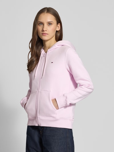 Vrouwenmodel draagt Regular Fit Sweatjacke aus Baumwoll-Mix van Tommy Jeans in lichtroze, capuchon met tunnelkoord, kangoeroezak, logo-stiksel, lange mouwen met geribde boorden, sweatstof van katoenmix, unie kleur, volledige rits, rechte fit. Vooraanzicht, bovenlichaam close-up.